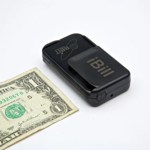 The iBill talking banknote identifier: a small matte black rectangular device, roughly the size of a matchbox, with a sliding slot cover on its top face printed with the word iBill, lying next to the corner of a one-dollar bill that shows its green Treasury seal and the numeral 1 — the bill is bigger than the device.